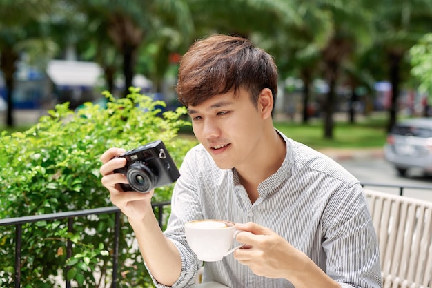 Portrait of good-looking guy holding photo camera while sitting at the table with cup of coffee