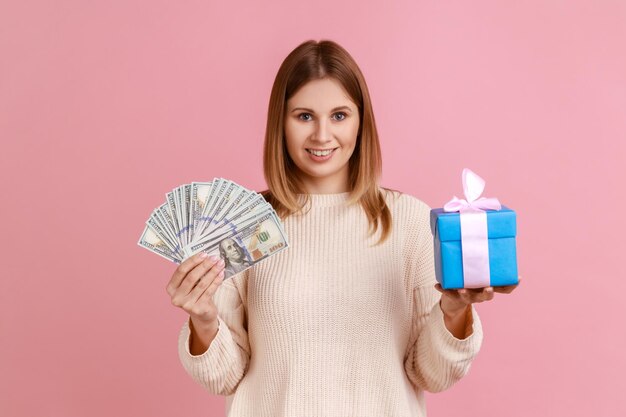 Portrait of good looking happy blond woman standing with wrapped present box and big fan of dollar banknotes in hands wearing white sweater Indoor studio shot isolated on pink background