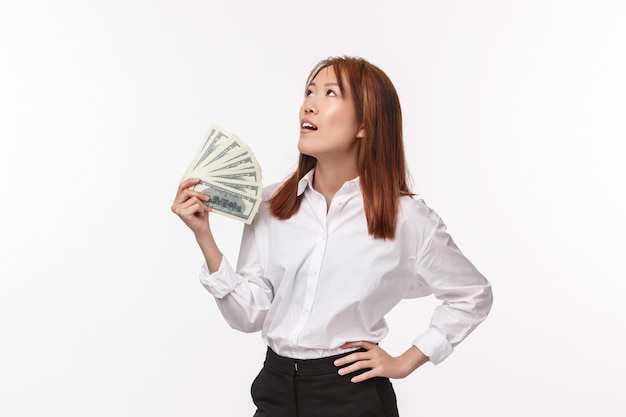 Portrait of good-looking successful asian woman in white shirt, businesswoman tired of being rich, waving money trying to cool herself look up relieved, become rich on white wall