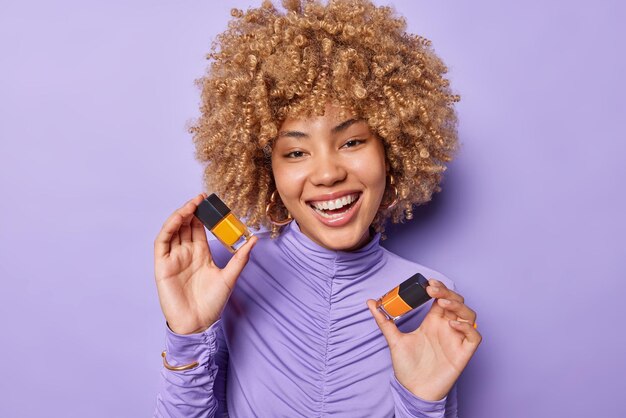 Portrait of good looking woman with curly hair holds two\
bottles of nail polish smiles positively wears golden earrings and\
turtleneck isolated over purple background beauty treatment and\
hand care