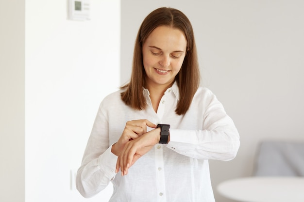 Portrait of good looking young adult woman wearing white casual style shirt, using her wrist watch, looking at smart phone screen, posing indoor in light room.