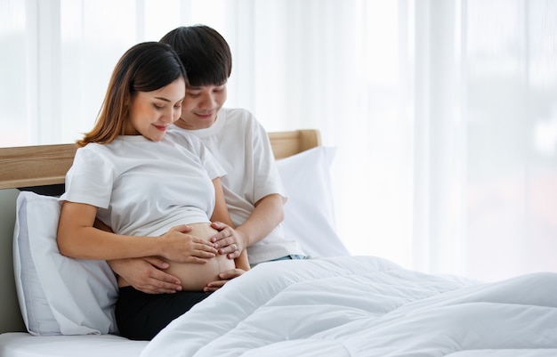 Portrait of a good-looking young Asian man and woman wearing a white nightdress sitting on a bed together. They are smiling happily and touching the belly of a pregnant mother.