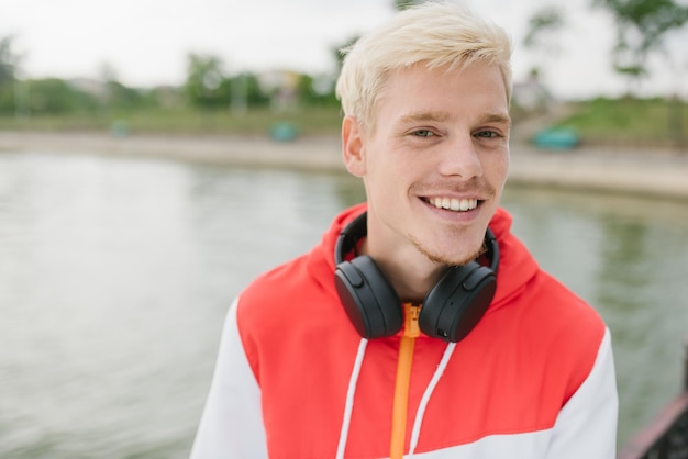 Portrait of goodlooking smiling attractive blond handsome man with headphones on neck walking and relax in the park of the city Street fashion style People lifestyle and technology concept