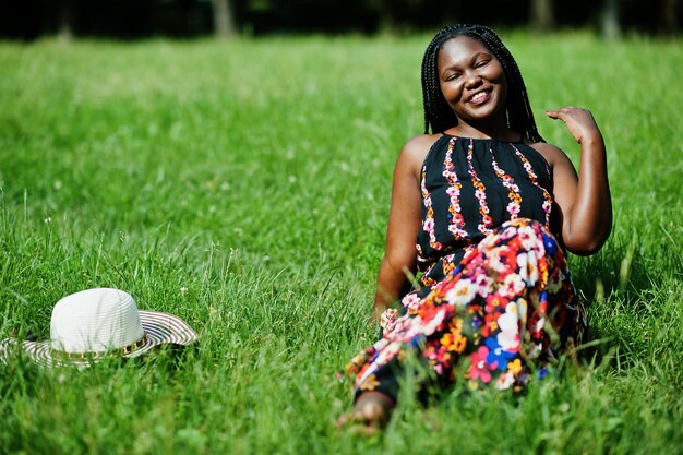 Portrait of gorgeous african american woman 20s in summer hat sitting at green grass in park.