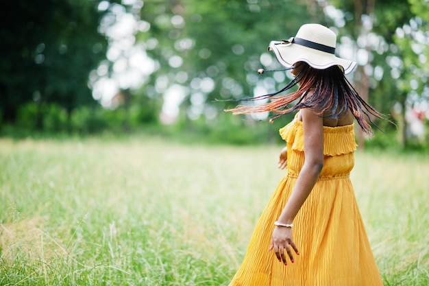 公園の緑の芝生でポーズをとって黄色のドレスと夏の帽子を身に着けているゴージャスなアフリカ系アメリカ人女性20代の肖像画。