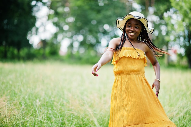 Portrait of gorgeous african american woman 20s in wear in yellow dress and summer hat posing at green grass in park