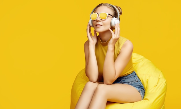 カラフルな黄色の背景にヘッドフォンで袋袋に座っているサングラスを身に着けているゴージャスな金髪の流行に敏感な女の子の肖像画