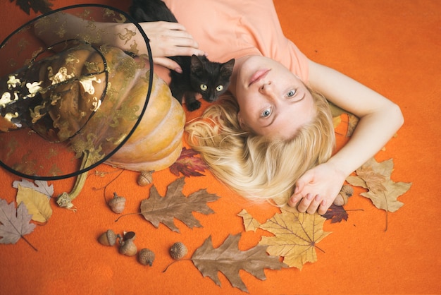 Portrait of gorgeous blonde witch and hat with pumpkin over orange background. Wide Halloween party art design. Beautiful young woman in black witch costume with hat over orange background.