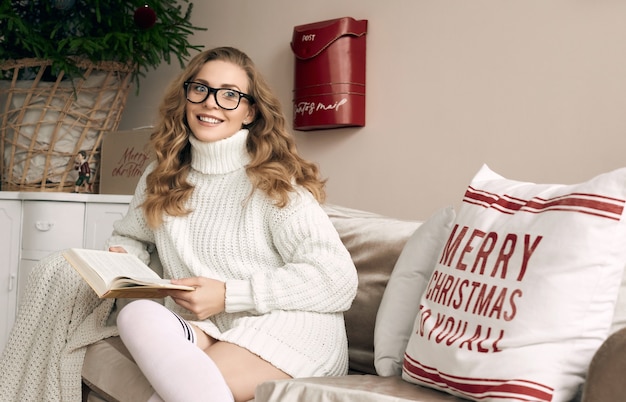 Portrait of gorgeous blonde woman in white woolen sweater and glasses reading book in light cozy decorated interior