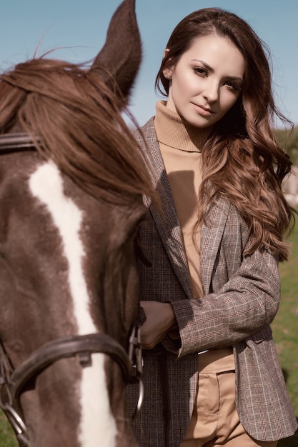 Portrait of a gorgeous brunette woman in an elegant checkered brown jacket posing with a horse on country landscape