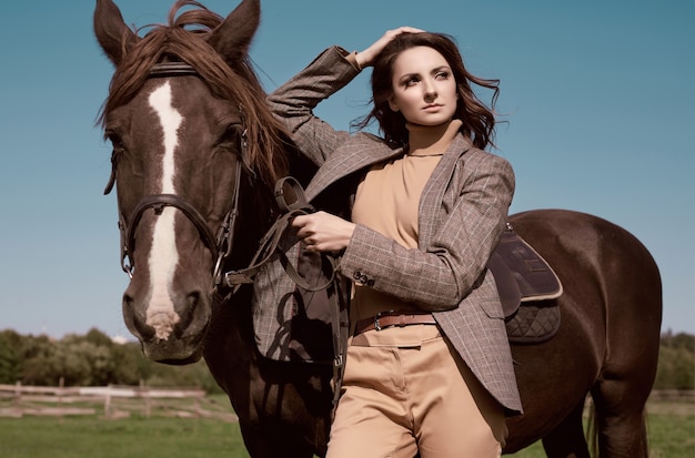 Портрет великолепной брюнетки в элегантной клетчатой коричневой куртке, позирующей с лошадью на деревенском пейзаже