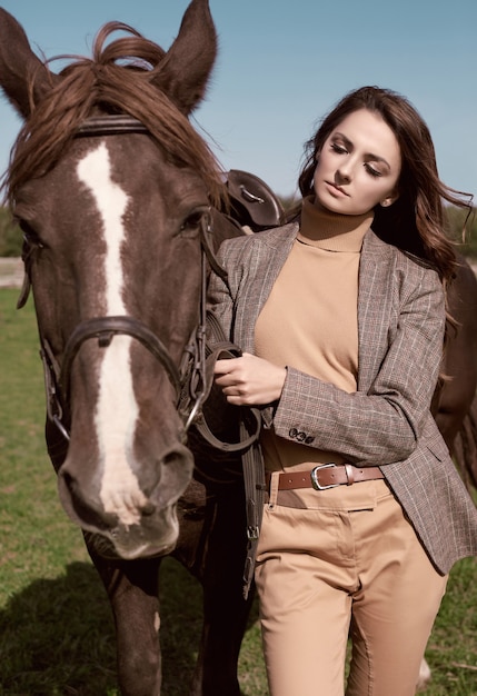 Portrait of a gorgeous brunette woman in an elegant checkered brown jacket posing with a horse on country landscape