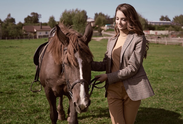 Ritratto di una splendida donna bruna in un'elegante giacca marrone a scacchi in posa con un cavallo sul paesaggio del paese