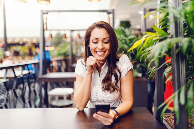 Portrait of gorgeous caucasian brunette in shirt using smart phone and sitting at bar.