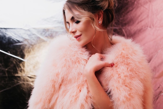 Portrait of gorgeous dreamy smiling girl in soft fluffy clothes posing with peacful face expression. Beautiful graceful young woman with elegant hairstyle standing in front of  sparkle wall