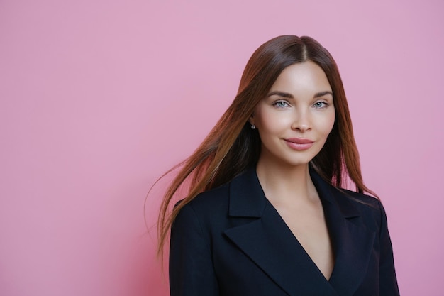 Portrait of gorgeous female manager going to work wears black formal costume natural makeup looks self confident at camera stands against pink studio background People and care concept