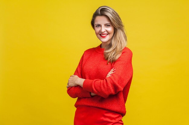 Portrait of gorgeous happy blonde woman with red lipstick in bright casual sweater standing with crossed arms and looking at camera with toothy smile indoor studio shot isolated on yellow background