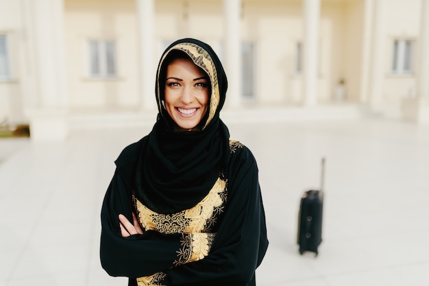 Portrait of gorgeous muslim woman with toothy smile and arms crossed standing. In background luggage.