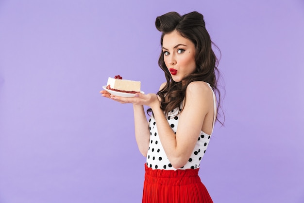 Portrait of gorgeous pin-up woman in vintage polka dot dress smiling while holding and eating sweet cheesecake isolated over violet wall