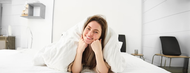 Photo portrait of gorgeous smiling woman lying in bed covered in duvet has messy hair looking happy