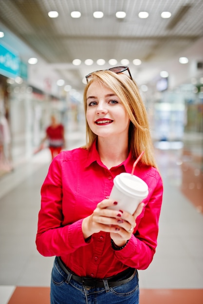 Portrait of a gorgeous woman in red blouse and jeans holding a cup of coffee in a shopping mall.