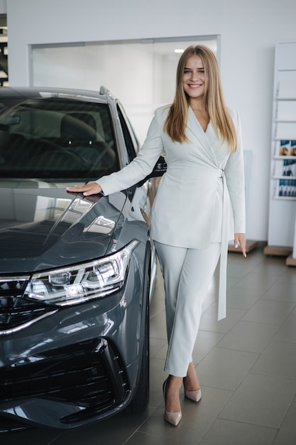 ショールームでゴージャスな女性の肖像画後の車のブロンドの髪の女性のそばに立っている美しい販売員