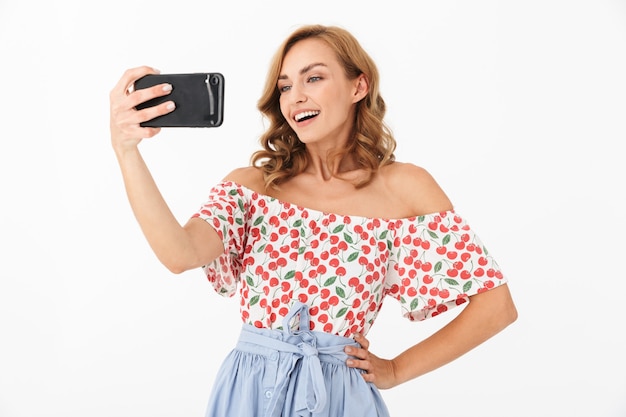 Portrait of gorgeous young woman dressed in summer clothes smiling while taking selfie photo on cellphone isolated