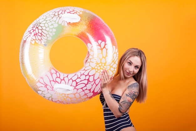 Portrait of gorgeous young woman looking at the camera and playing with colorful swimming ring in studio over yellow background. Pretty blonde in blue swimming suit.
