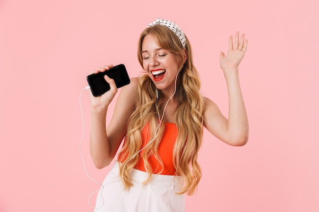 Портрет великолепной молодой женщины с длинными вьющимися волосами поет, держа смартфон с наушниками, изолированными над розовой стеной