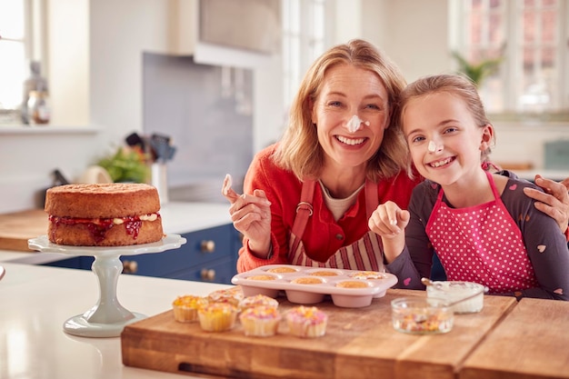 Portrait Of Grandmother With Granddaughter Baking Cupcakes And Putting Icing On Their Noses At Home