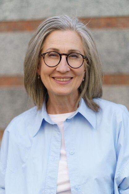 Portrait of grey hair mature woman standing outdoors on marble wall background smiling looking at camera Standing outdoors positive gorgeous woman in casual blue shirt