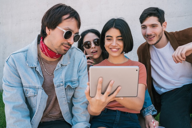 Portrait of group of friends having fun and using a digital tablet together while sitting outdoors. Technology, lifestyle and friendship concept.