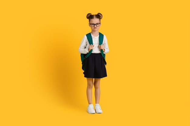 Portrait of grumpy little schoolgirl with backpack standing over yellow studio background