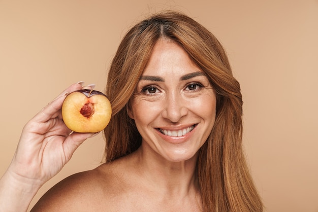 Portrait of half-naked adult woman holding plum and smiling isolated on beige