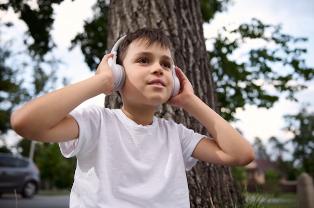 Portrait of handsome adorable child boy, wearing wireless headphones and enjoying listening to music, resting during recreation between classes in the public city park, after his first day at school