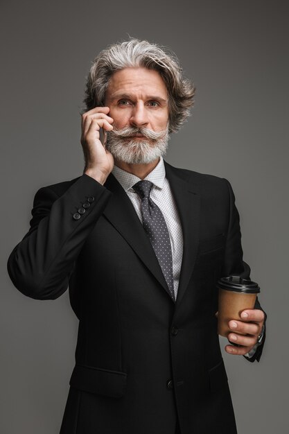 Portrait of handsome adult businessman wearing formal black suit holding paper cup and talking on smartphone isolated over gray wall