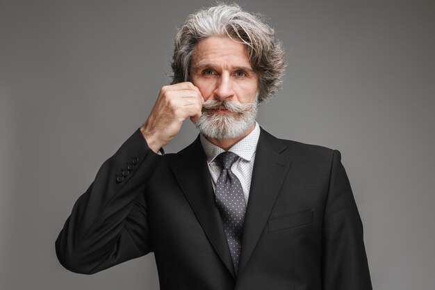 Portrait of handsome adult businessman wearing formal black suit looking at front and touching his mustache isolated over gray wall