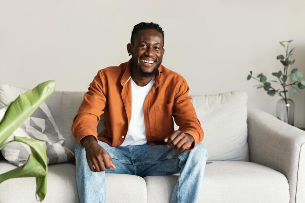 Portrait of handsome african american man smiling and looking at camera sitting on sofa at home free space