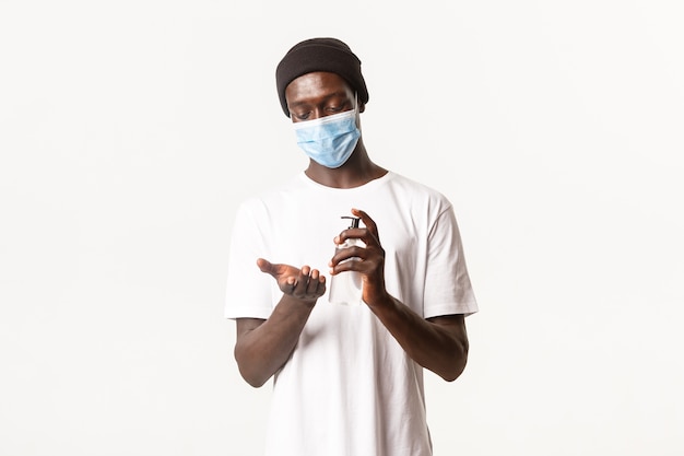 Portrait of handsome african-american young guy in medical mask applying antiseptic on hands