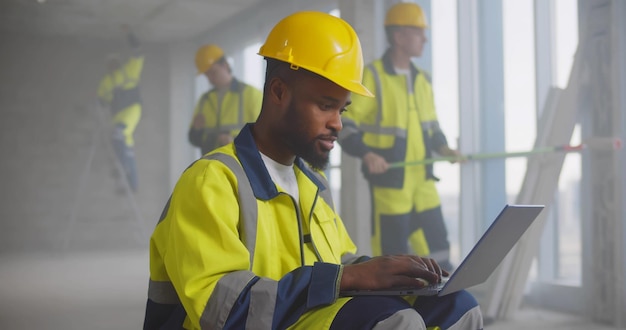 Portrait of handsome AfricanAmerican builder in helmet sitting with laptop at construction site