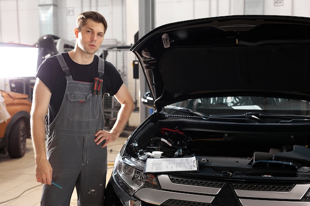 Portrait of handsome auto mechanic man standing near the car hood in auto service workshop