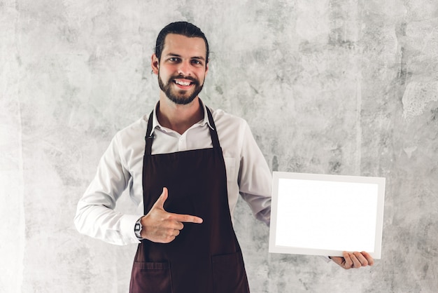 Portrait of handsome bearded barista man small business owner smiling and holding empty board wooden frame with white  blank in a cafe