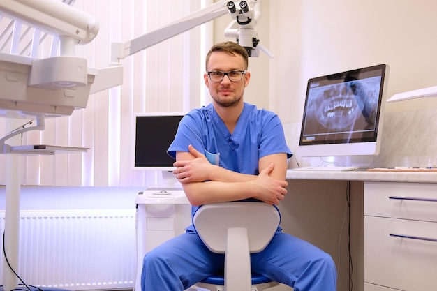 Portrait of a handsome bearded dentist male in glasses wearing a blue uniform, sitting in a dentist office. Looking at camera.