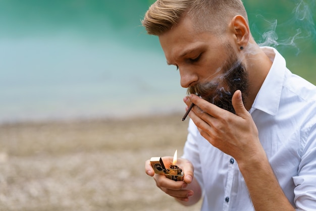 Photo portrait of a handsome bearded guy lights a cigarette with a lighter outdoors on the background of a...