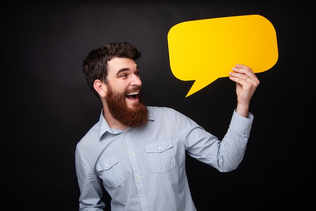 Portrait of handsome bearded guy with beard, excited holding a yellow empty bubble speech, standing over dark isolated background