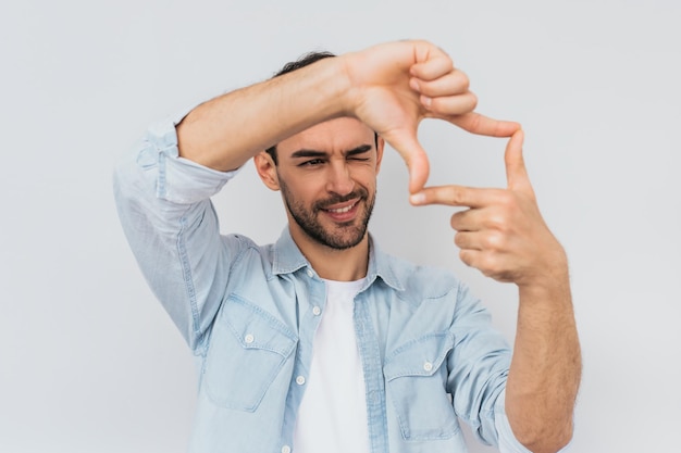 Portrait of handsome bearded happy male is looking through a frame formed by his hands Attractive man make frame by fingers isolated on a white background with copy space for text People concept