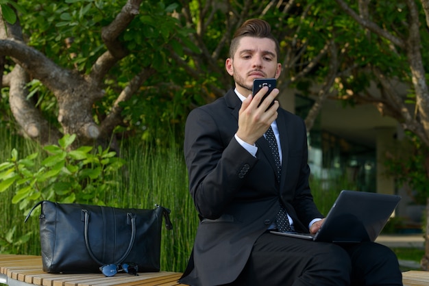 Portrait of handsome bearded Hispanic businessman with nature in the city outdoors