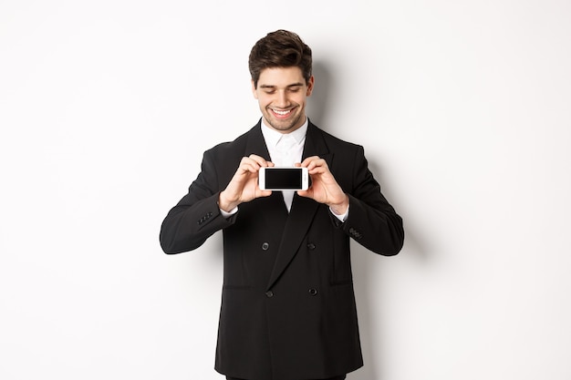 Portrait of handsome bearded man in black suit, showing you smartphone screen and smiling pleased, standing against white background.