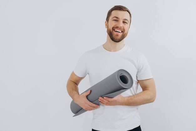 Portrait of handsome bearded man in sportswear holding yoga mat on white background copy space