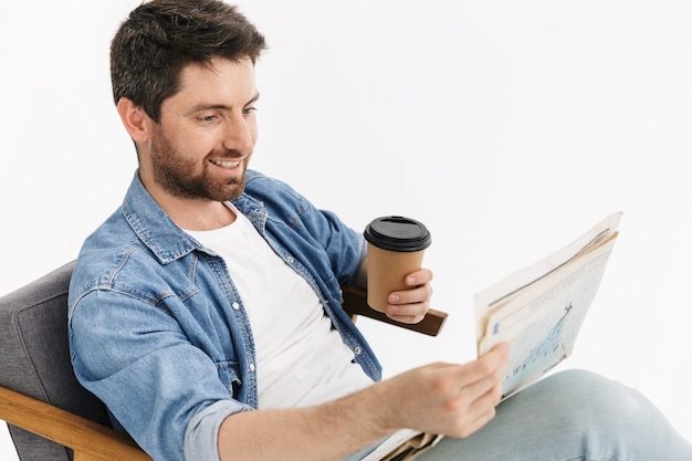 Portrait of a handsome bearded man wearing casual clothes sitting in chair isolated, reading newspaper, drinking coffee